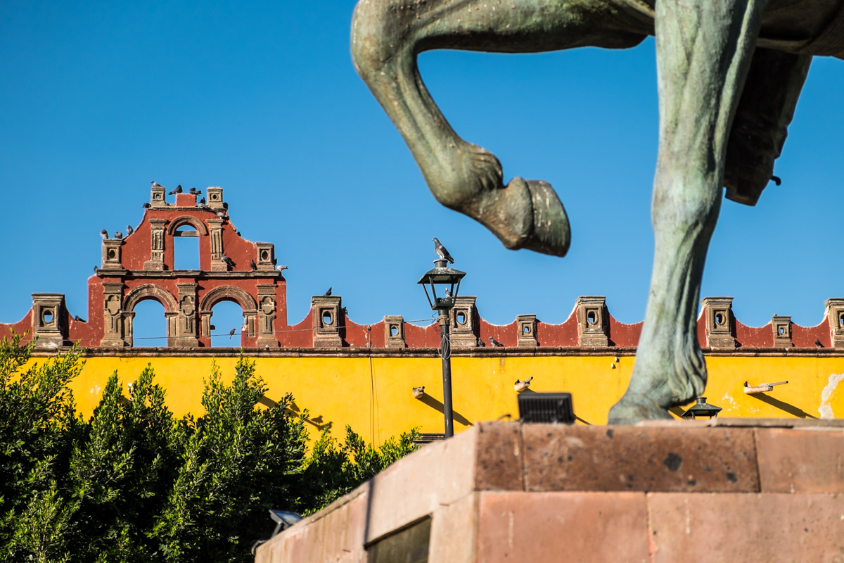 In a colorful square in San Miguel Allende, a statue of a horse appears to be trying to kick a pigeon perched on a lamppost.